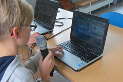Ein Schüler sitzt vor einem Laptop und hält ein Mikrophone in der Hand
